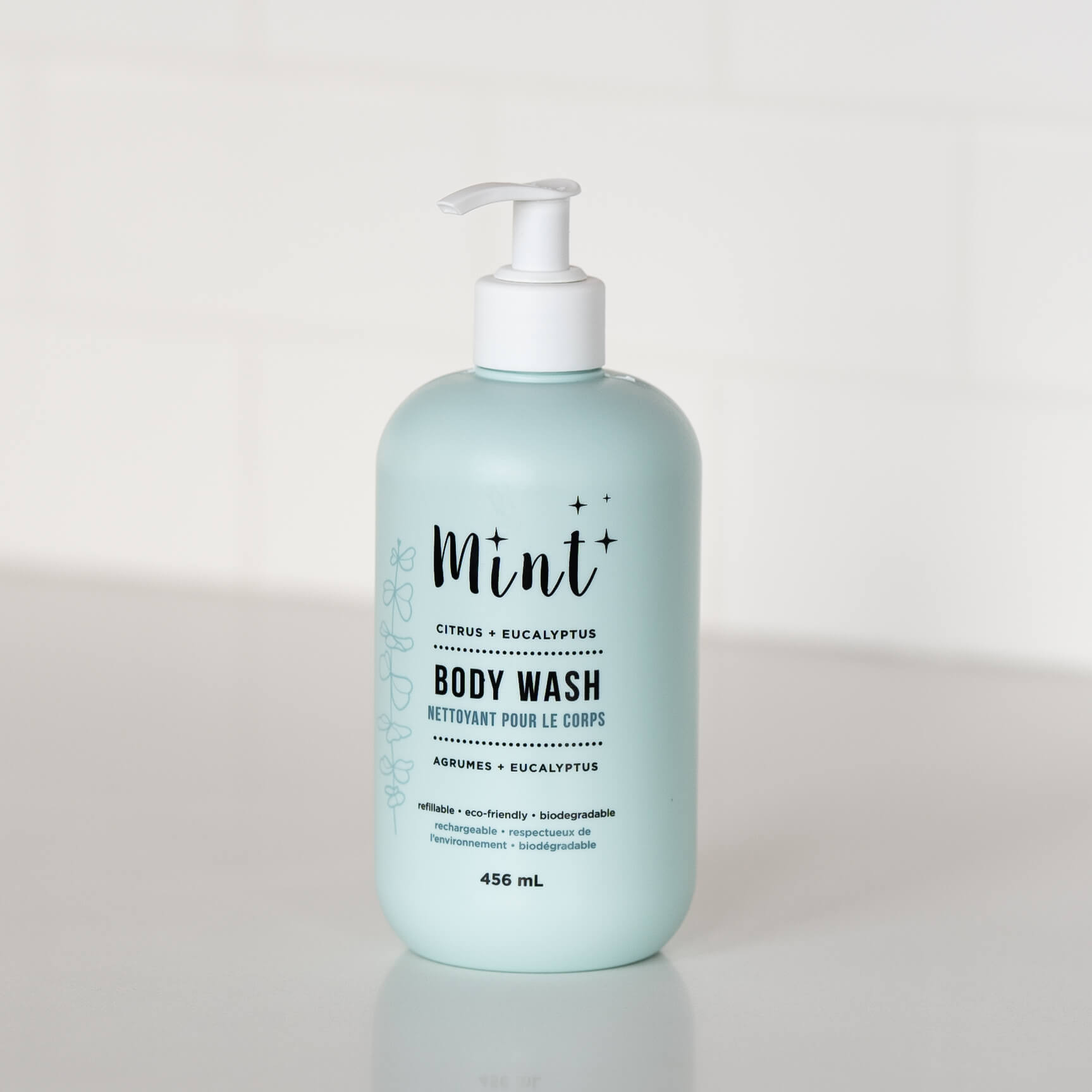 A 456 mL light blue bottle of "Mint+ Body Wash" with a pump dispenser, displayed on a white countertop. The label features the scents "Citrus + Eucalyptus" with bilingual text "Nettoyant pour le corps" and highlights eco-friendly attributes such as being refillable, biodegradable, and sustainable. Subtle botanical illustrations adorn the left side of the label, creating a clean and natural presentation. The background includes a soft, minimalistic white setting.