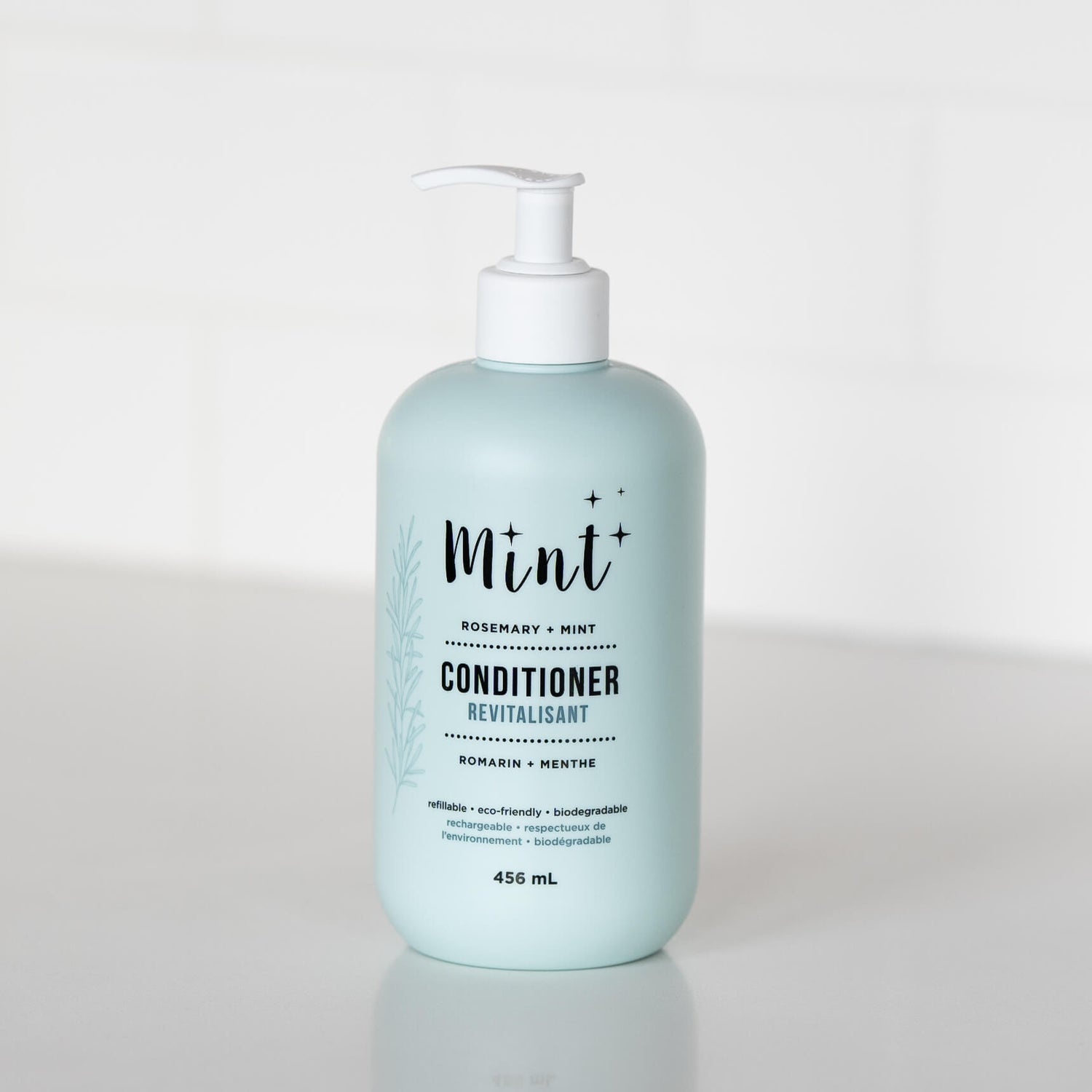 A 456 mL light blue bottle of &quot;Mint+ Conditioner&quot; with a pump dispenser, displayed on a white countertop. The label highlights the scent &quot;Rosemary + Mint&quot; and includes bilingual text, &quot;Revitalisant&quot; and &quot;Romarin + Menthe.&quot; It emphasizes eco-friendly attributes such as being refillable, biodegradable, and sustainable. A subtle botanical illustration adorns the left side of the label, adding a natural element to the design. The background features a bright and minimalistic white setting.