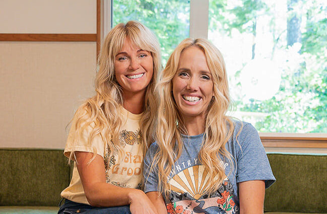 Monika and Robyn, co-founders of Mint Cleaning Products, sitting together on a green couch, smiling at the camera. Monika is wearing a yellow graphic t-shirt, and Robyn is in a blue graphic t-shirt. A large window behind them lets in natural light, with greenery visible outside