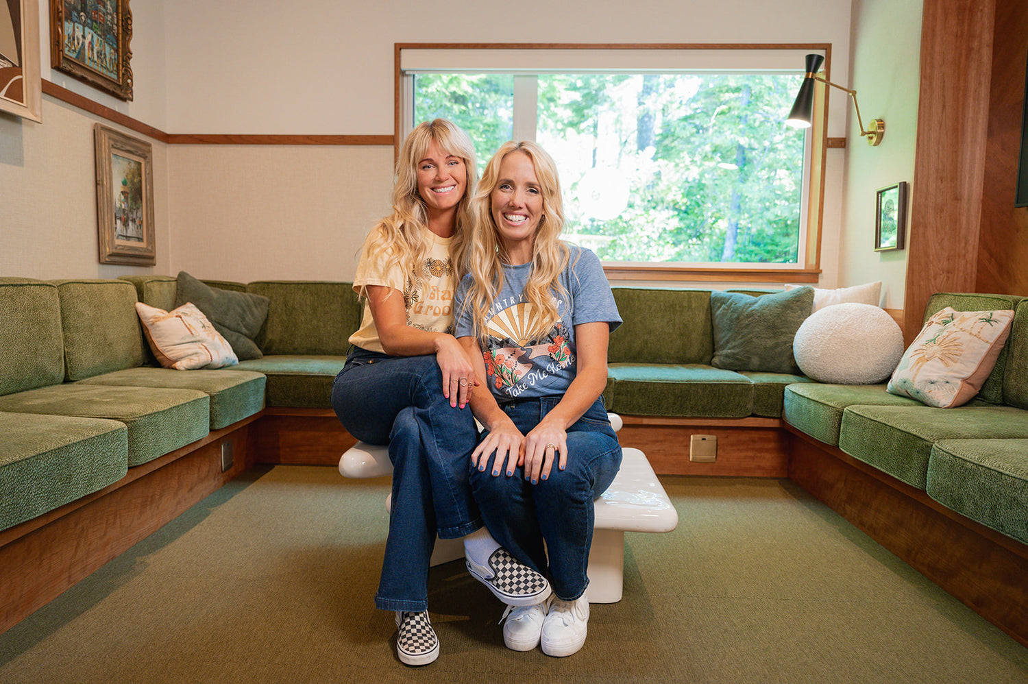 Monika and Robyn, co-founders of Mint Cleaning Products, sitting together on a green couch, smiling at the camera. Monika is wearing a yellow graphic t-shirt, and Robyn is in a blue graphic t-shirt. A large window behind them lets in natural light, with greenery visible outside