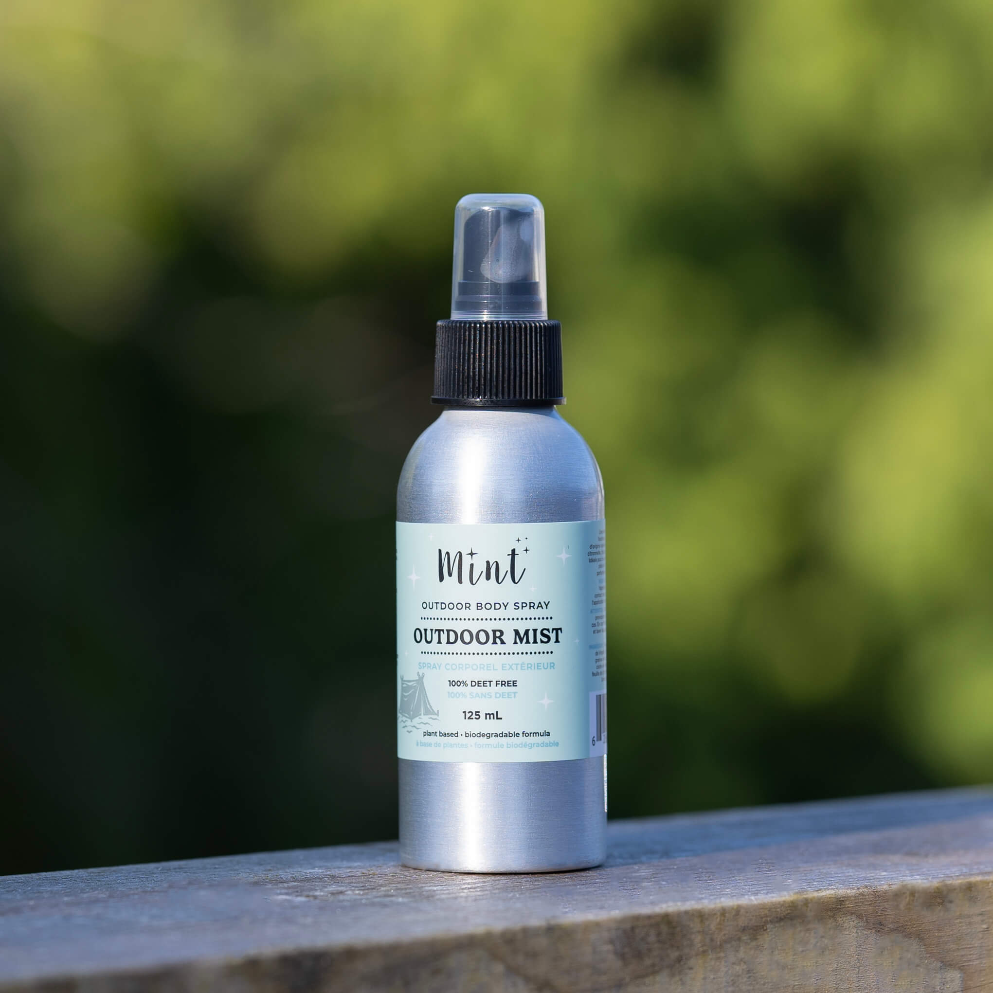 Mint Outdoor Mist body spray bottle placed on a wooden railing outdoors, with a blurred green natural background. The aluminum bottle features a mint green label and black spray cap, emphasizing its eco-friendly and plant-based formula.