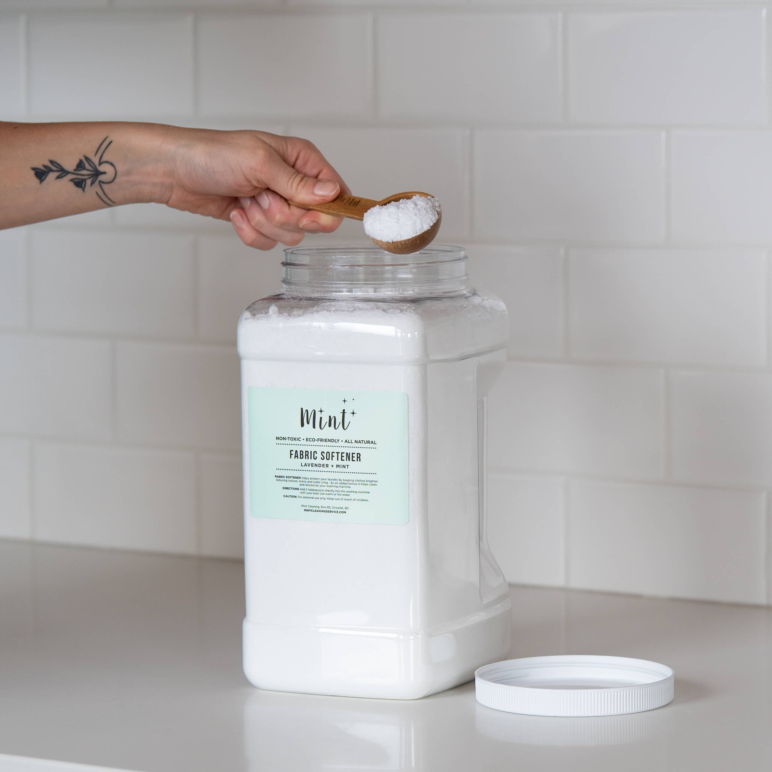 A large white container of "Mint+ Fabric Softener - Lavender + Mint" with its lid off, placed on a white countertop. A hand with a tattoo is holding a bamboo scoop with product over the open container, against a minimalist subway tile background.