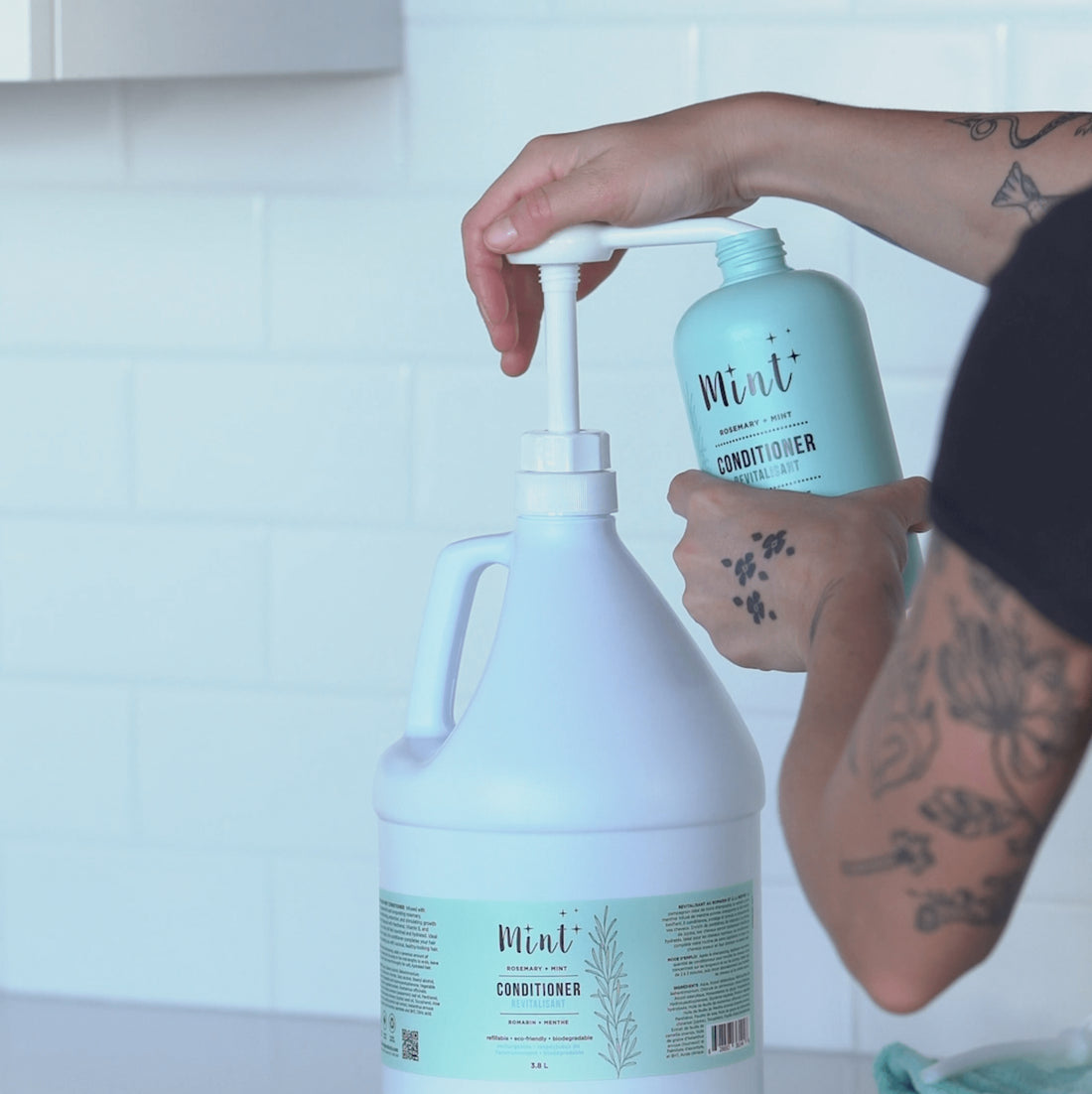 A person refilling a smaller bottle of Mint Rosemary + Mint Conditioner from a large 3L bulk container. The container is white with a label featuring the Mint logo and product details, and the person’s hands are holding both the pump of the large container and the smaller bottle. The background features a clean, white tiled wall.