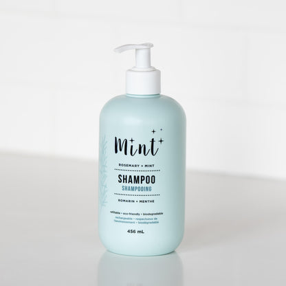 A 456 mL light blue bottle of &quot;Mint+ Shampoo&quot; with a pump dispenser, displayed on a white countertop. The label highlights the scent &quot;Rosemary + Mint&quot; and includes bilingual text, &quot;Shampooing&quot; and &quot;Romarin + Menthe.&quot; The product&