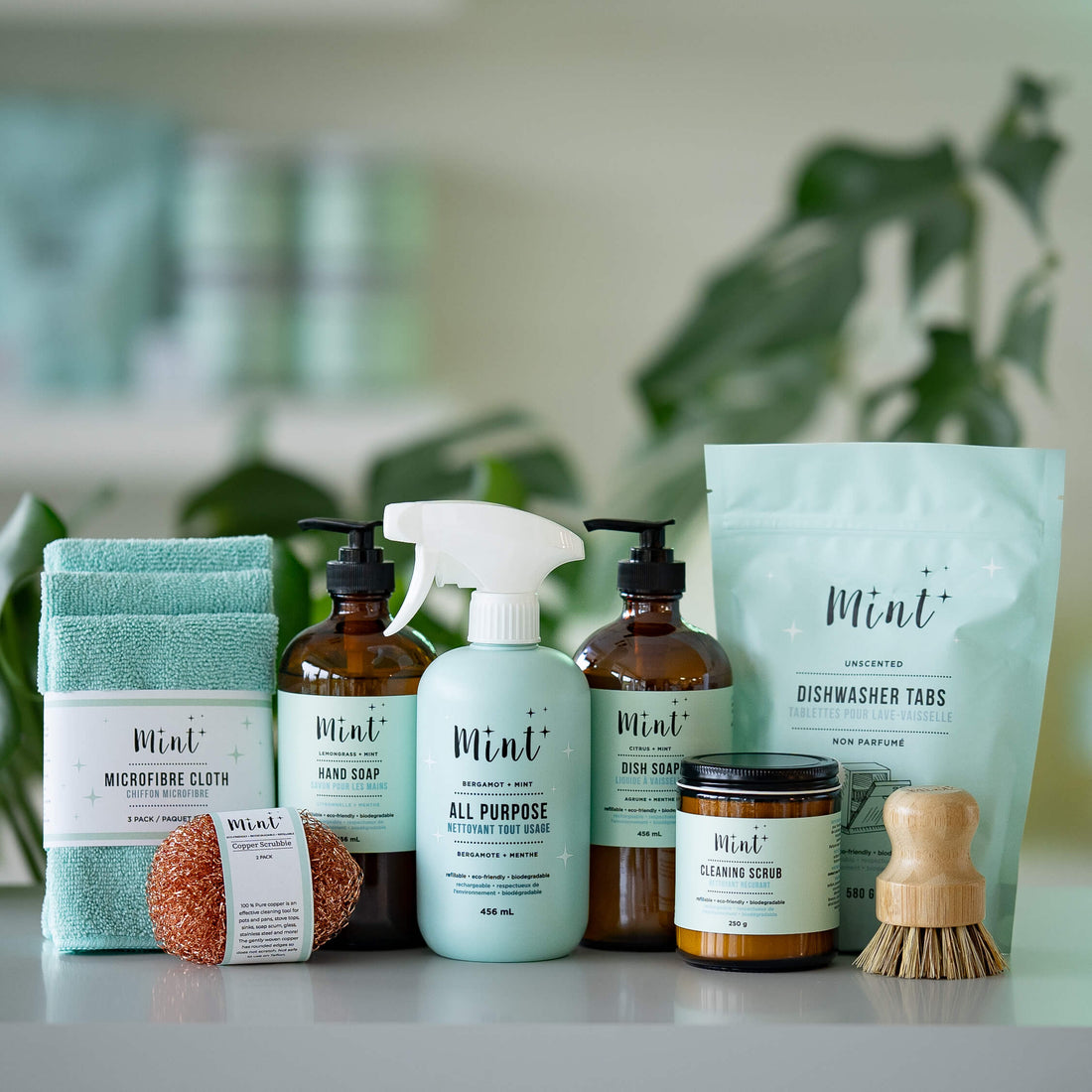 The Essential Kitchen Bundle by Mint Cleaning Products, featuring hand soap, dish soap, all-purpose cleaner, dishwasher tabs, cleaning scrub, microfiber cloths, a copper scrubber, and a wooden dish brush, displayed with green plants in the background