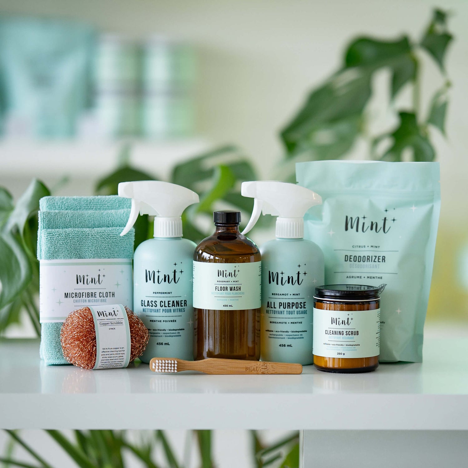The Ultimate Cleaning Bundle by Mint Cleaning Products, featuring glass cleaner, all-purpose cleaner, floor wash, deodorizer, cleaning scrub, microfiber cloths, a copper scrubber, and a bamboo cleaning brush, displayed with green plants in the background